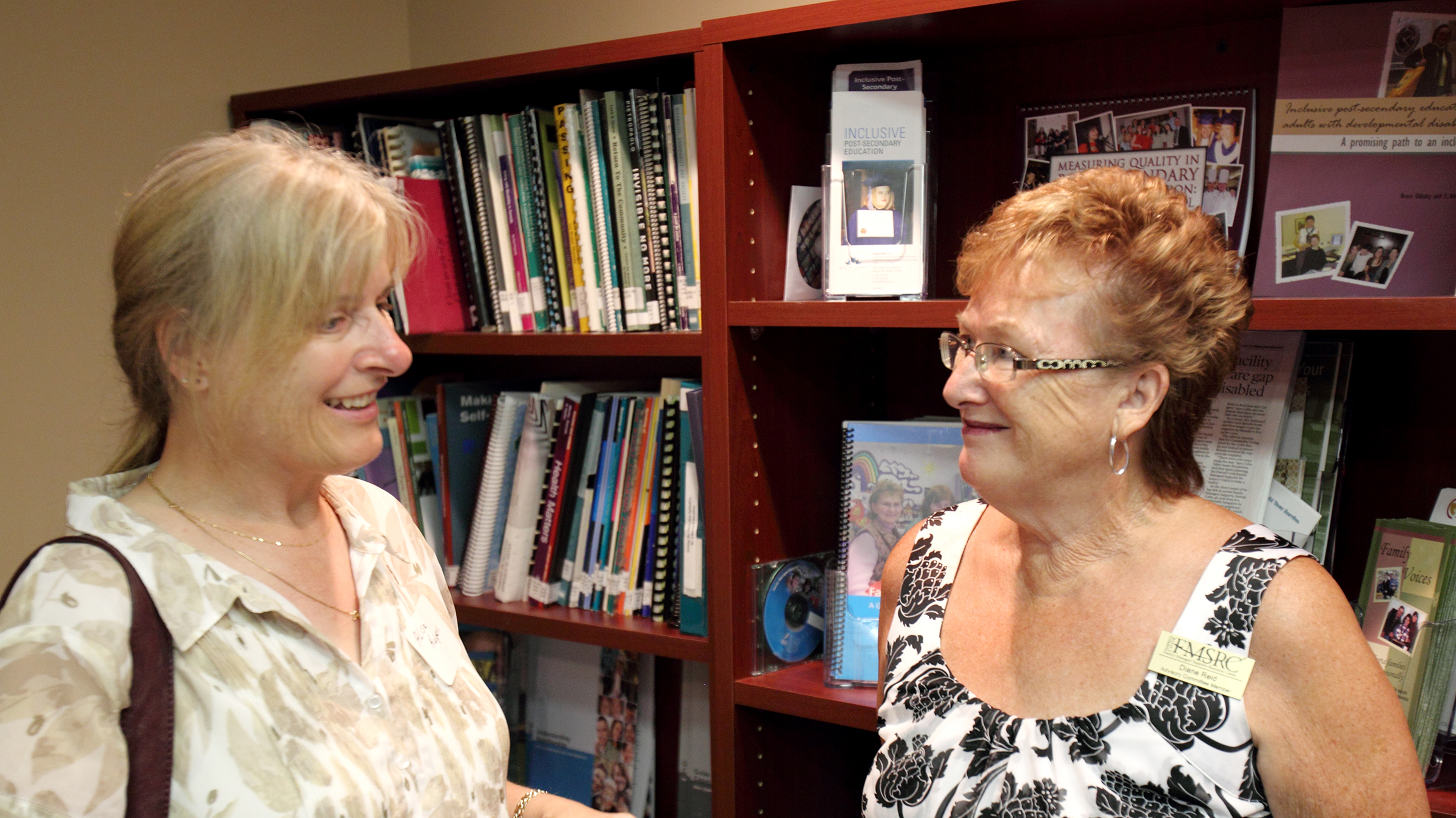 Two women smile while talking in front of full bookshelves